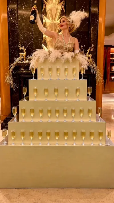 A performer in a gold beaded dress with a feather headpiece raising a champagne bottle above a tiered gold champagne tower display at a formal event