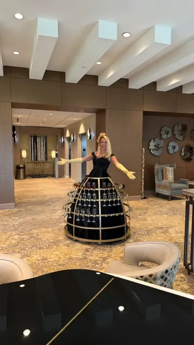 A roaming performer in a black dress with gold gloves and a circular champagne skirt display in a contemporary hotel lounge with a piano in the foreground
