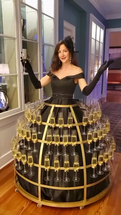 A roaming performer in a black off-the-shoulder dress with long gloves and a circular champagne skirt display, standing beside tall windows on a wooden floor